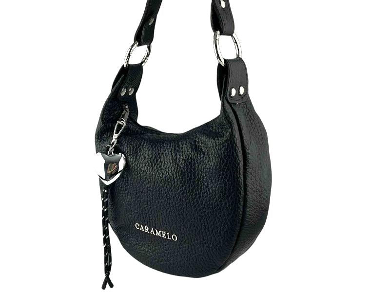 Vista lateral de un elegante bolso de mujer, realizado en piel de color negra, de diseño elegante y con líneas puras y minimalista, ideal con cualquier estilo. Con detalle de llavero extraíble, con forma de corazón plateado y cordones negros con detalles sutiles en blanco.