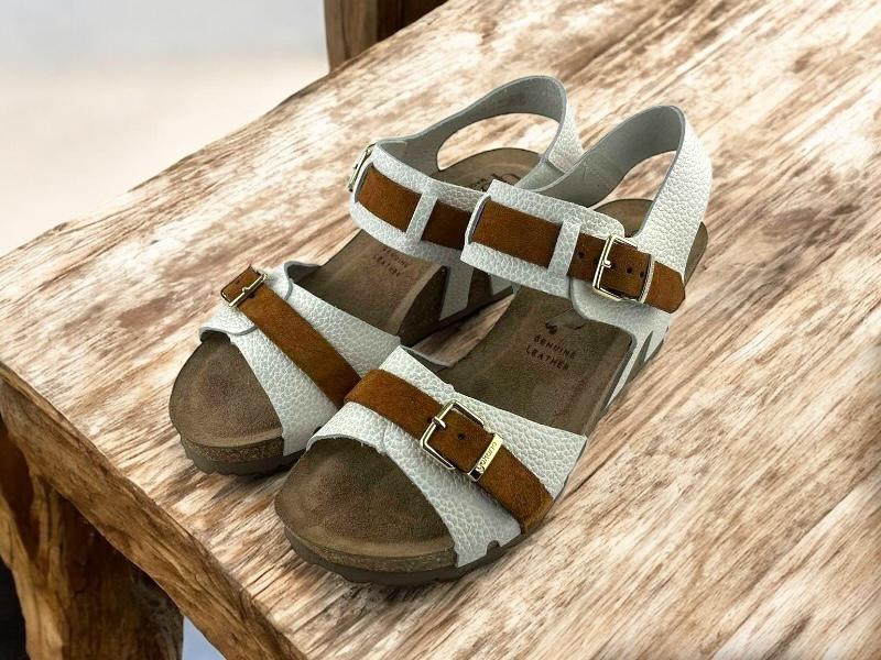 Vista lateral derecha, de unas sandalias en piel, de mujer, blancas, ergonómicas y con cuña. Cierre regulable por hebilla en tiras color avellana en empeine y tobillo sobre fondo de madera blanca.