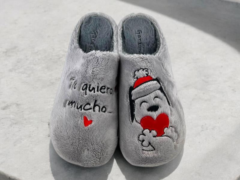 Zapatillas de casa, descalzas, mujer, paño grueso, color gris, con bordado en una zapatilla Te quiero mucho y en la otra un perrito con corazón rojo. Vista frontal de las zapatillas sobre fondo de mármol blanco.