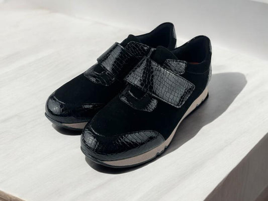 Tenis de piel, de mujer, en negro, con detalles en charol en la tapeta y en el velcro. Vista lateral izquierda sobre fondo de madera blanca.