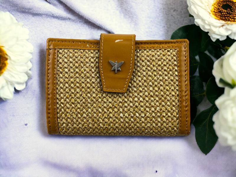 Cartera de mujer en piel color mostaza y rafia beige. Logo de la marca en la solapa. Vista frontal por la parte de atrás sobre fondo de flores.