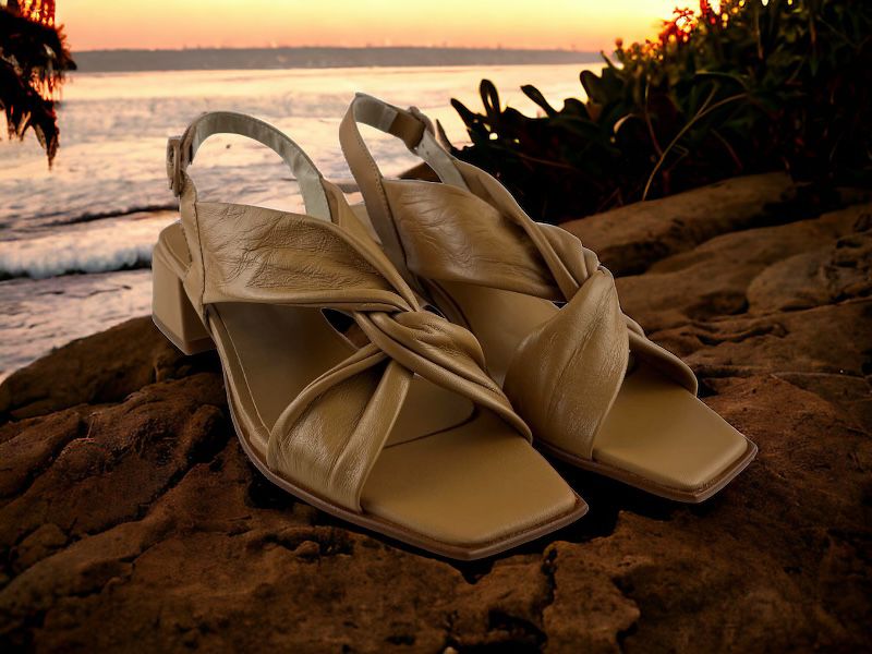 Sandalias de piel color cuero con nudo en el empeine, puntera cuadrada y tacón geómétrico. Vista lateral derecha de las sandalias sobre fondo de arena en el atardecer de una playa.