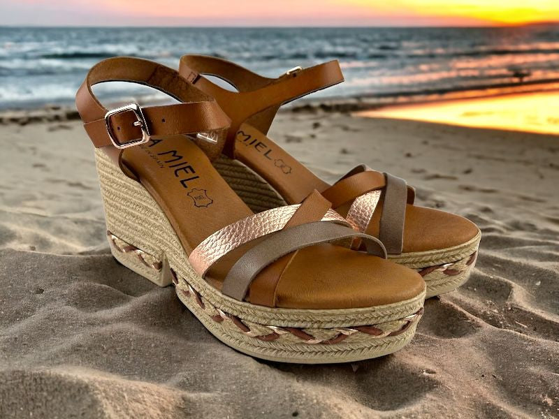 Sandalias de mujer en piel, con pala en colores metalizados, con cuña de esparto. Vista lateral derecha sobre fondo de arena de playa.