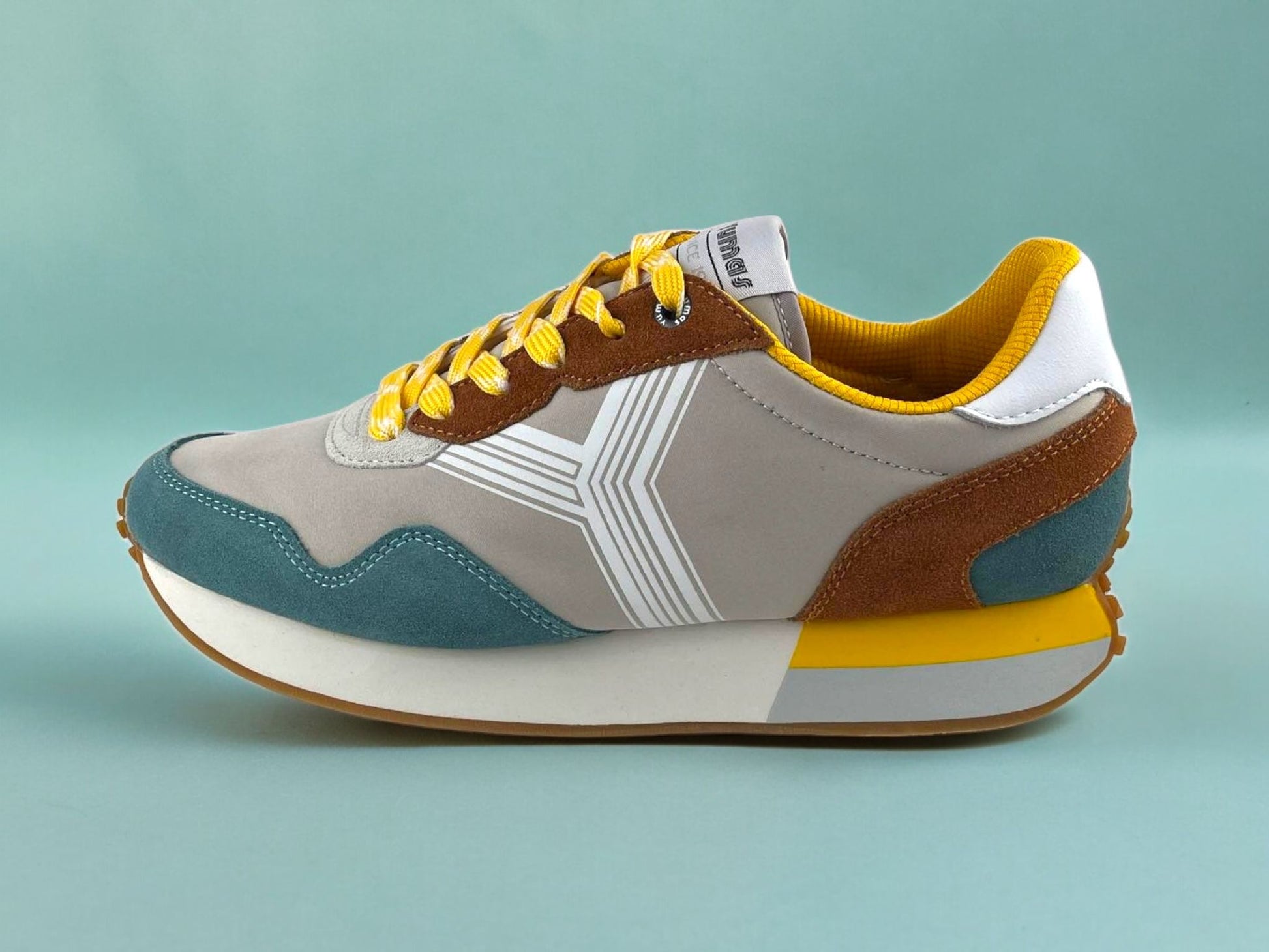 Tenis de hombre en beige, con logo en blanco, puntera y talonera en azul y ocre. Suela en blanco, azul y amarillo. Vista lateral izquierda sobre fondo azul.