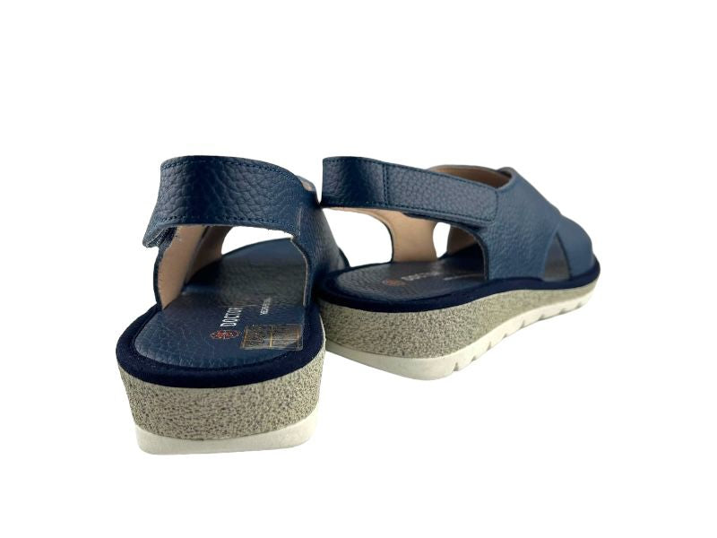 Sandalias de mujer en color azul marino con tiras cruzadas en empeine y cierre de Velcro al tobillo. Cuña en color gris y suela blanca. Vista  por la parte de atrás sobre fondo blanco.