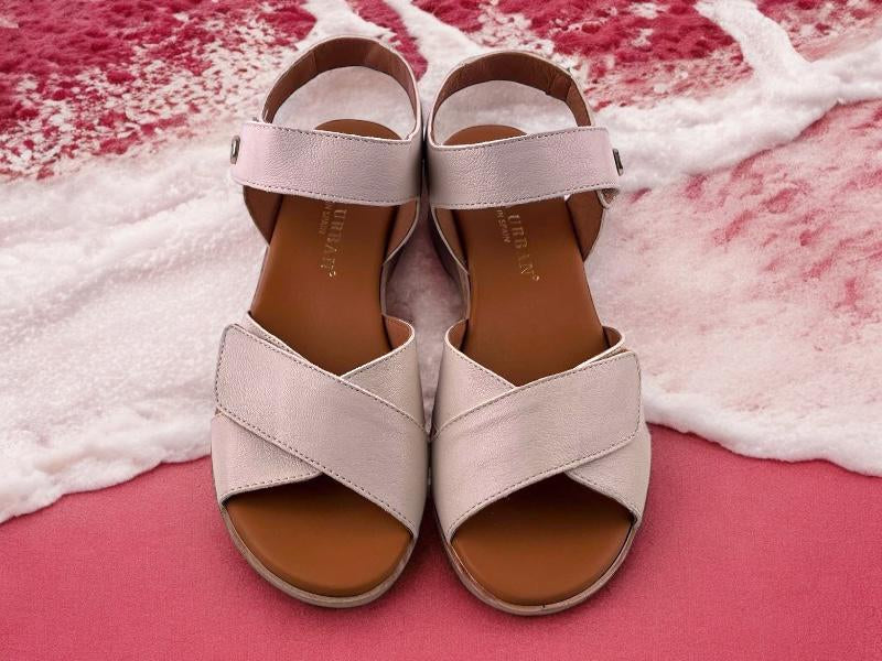 Sandalias planas de mujer con tiras regulables cruzadas en color beige y pulsera en el tobillo. Vista frontal de las sandalias sobre espuma de mar en tono fucsia.