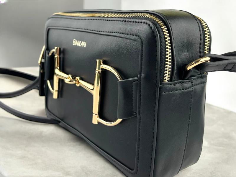 Bolso estilo bandolera negro, de la marca Binnari, con hebillas doradas y logo de la marca impreso. Vista delantera lateral izquierda  sobre fondo de mármol blanco.