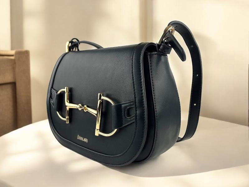 Bolso estilo bandolera  con solapa negro, de la marca Binnari, con hebillas doradas y logo de la marca impreso. Vista delantera lateral izquierda  sobre una mesa blanca.