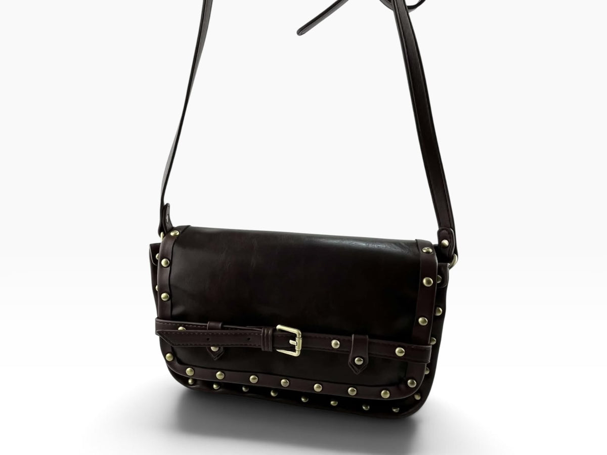 Bolso bandolera Pepe Moll de piel vegana marrón oscuro con solapa frontal, hebilla central y tachuelas doradas.