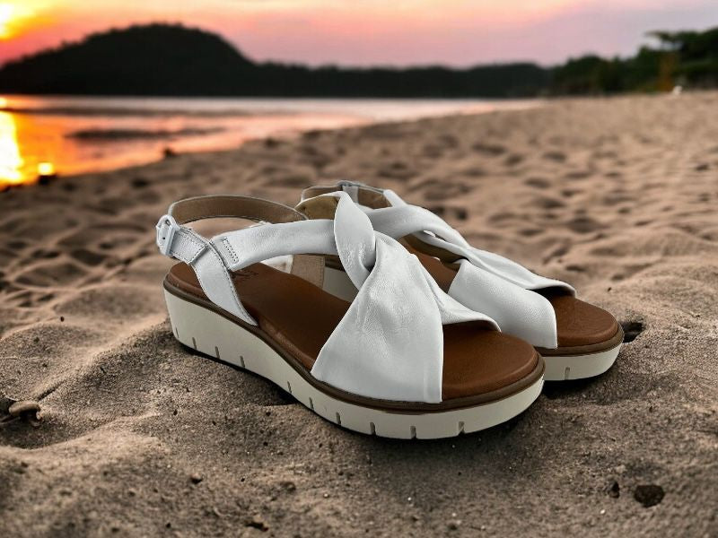 Sandalias de mujer en piel blanca, con nudo en el empeine y cierre con hebilla. Suela en beige claro. Vista lateral derecha sobre fondo de arena.