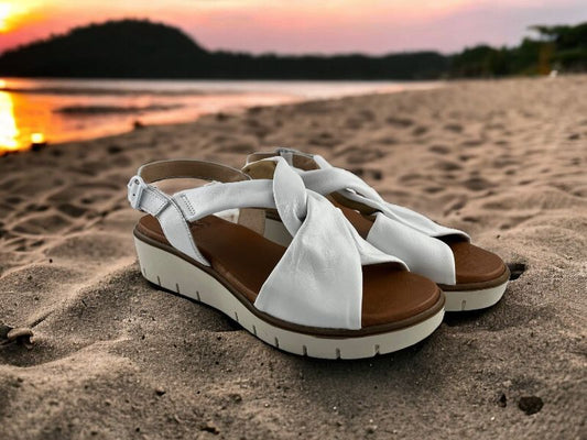Sandalias de mujer en piel blanca, con nudo en el empeine y cierre con hebilla. Suela en beige claro. Vista lateral derecha sobre fondo de arena.