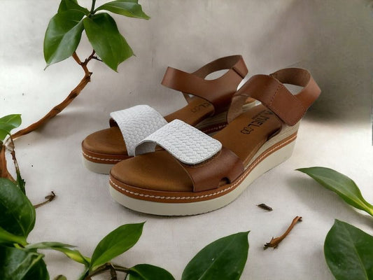 Sandalias de mujer en piel, con cuña, en color cuero y blanco. Vista lateral izquierda sobre fondo floral.