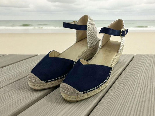 Vista lateral de un par de alpargatas artesanales de cuña baja. La parte delantera es de piel de ante azul marino con escote en V. La suela y la puntra reforzaeda son de yute trenzado color natural. Con pulsera de ante azul con hebilla dorada ajustable.