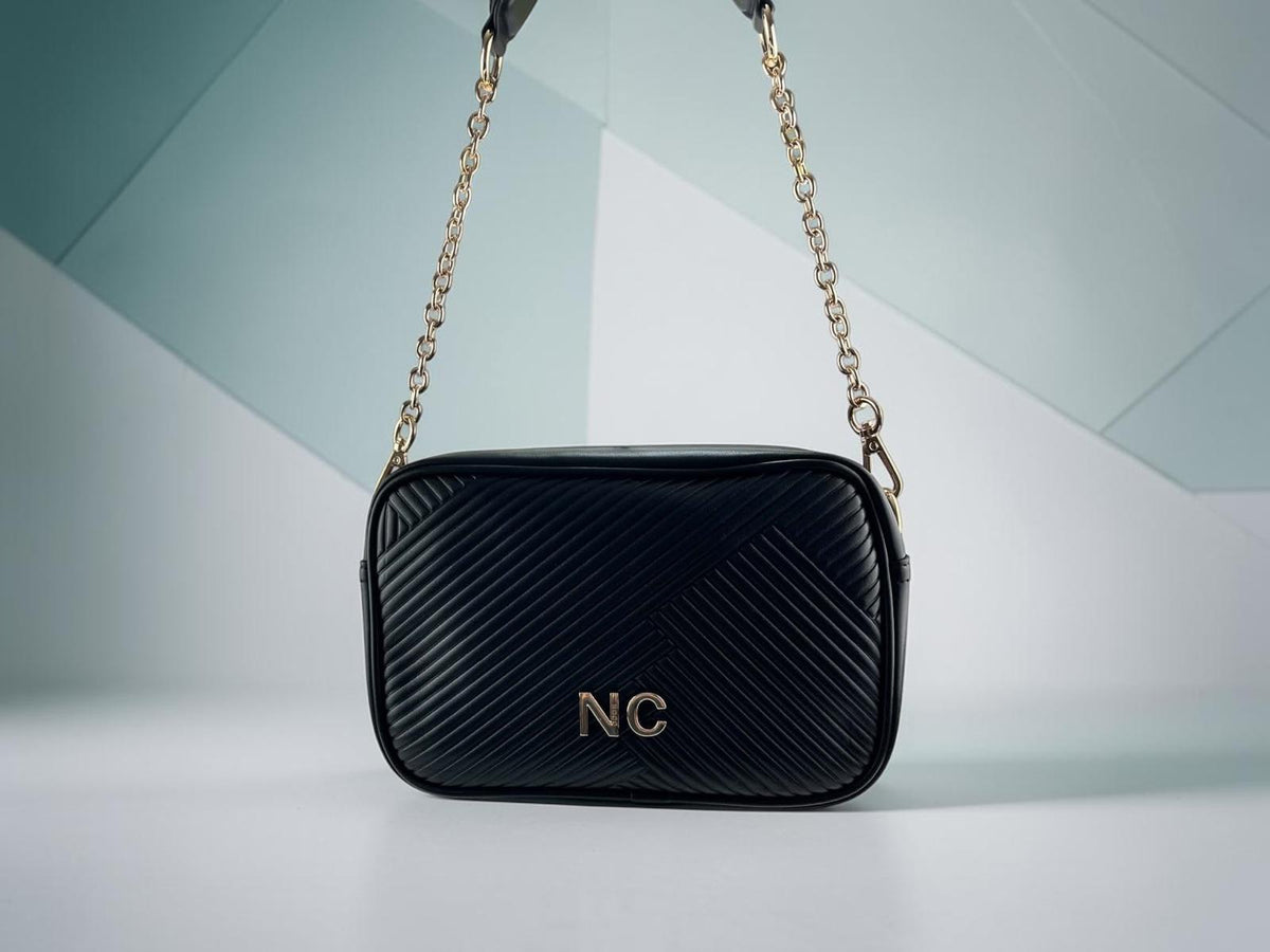 Bandolera Noco negra de piel vegana con textura plisada diagonal, cadena dorada y detalle NC, vista frontal.