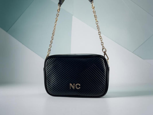 Bandolera Noco negra de piel vegana con textura plisada diagonal, cadena dorada y detalle NC, vista frontal.