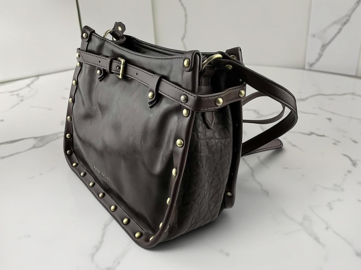 Bolso bandolera Pepe Moll de piel vegana marrón oscuro con tachuelas metálicas, hebilla decorativa y vista lateral.