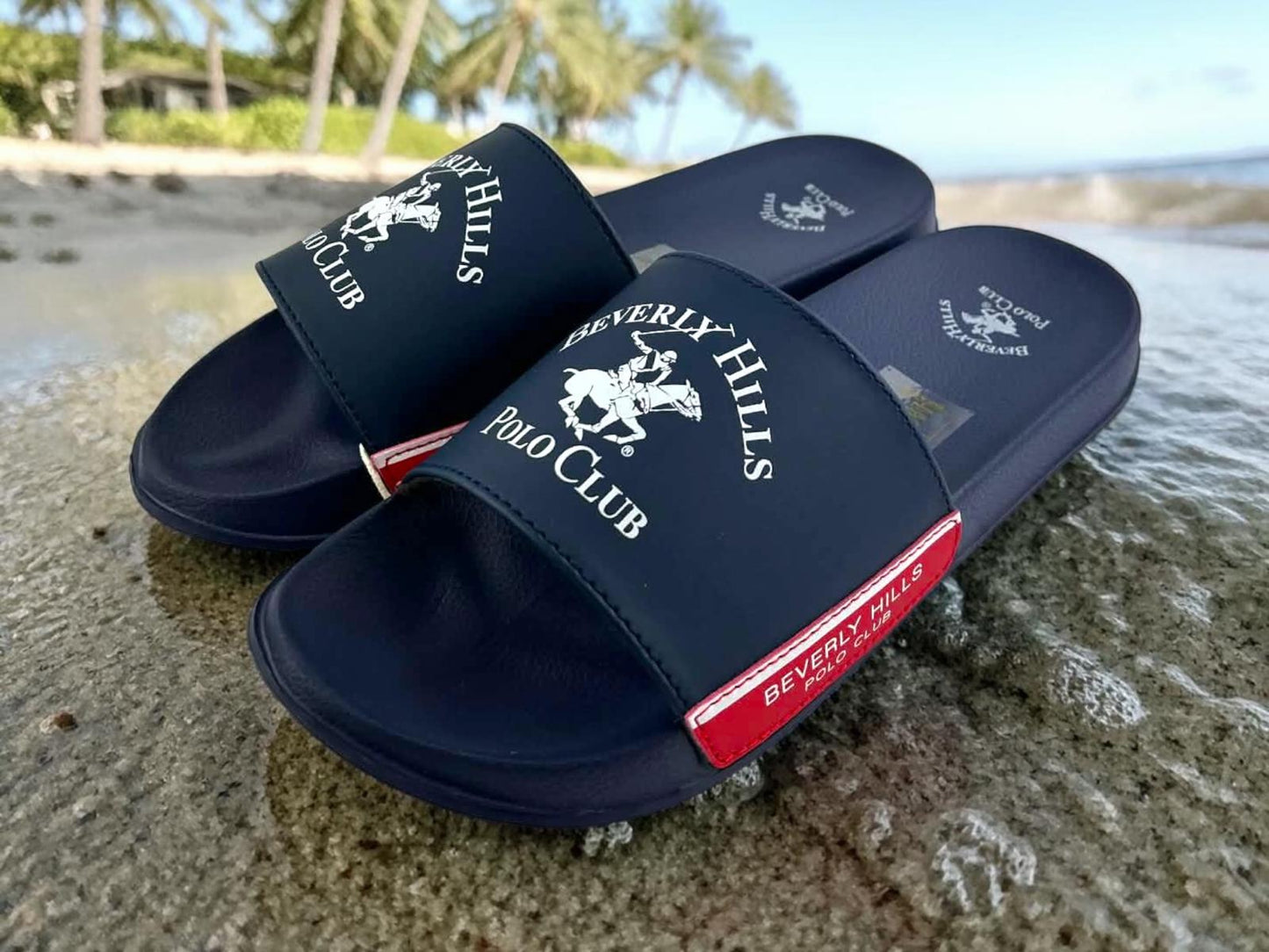 Chanclas de pala Beverly Hills Polo Club en color azul marino con logo blanco y detalle lateral en rojo deportivo sobre la arena mojada en una playa.