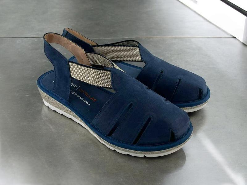 Vista lateral  derecha de unas sandalias planas en piel azul, de cortes asimétricos, con elásticos en el empeine y puntera cerrada sobre fondo gris.