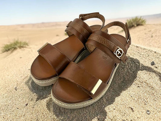 Sandalias de cuero marrón sobre arena de playa, resaltando su suela de plataforma resistente y diseño de pala con velcro para máxima comodidad en exteriores.