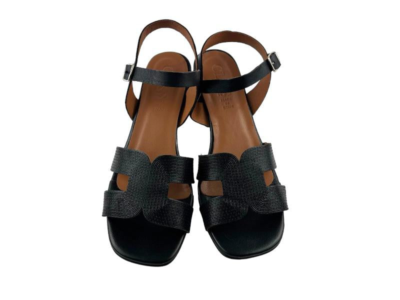 Sandalias de mujer con pala geométrica y troquelada negra; cierre de hebilla y tacón medio geométrico forrado en negro. Vista lateral frontal sobre fondo color blanco.
