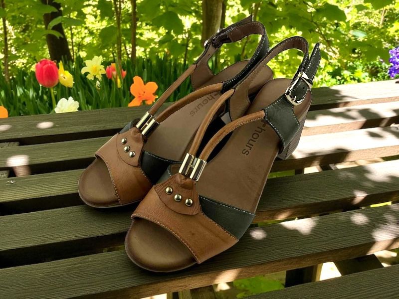 Sandalias de piel  de mujer bicolor color cuero y verde oliva, con tiras de cuero en el empeine y abotonadura al tobillo. Fondo de madera verde y flores de primavera.