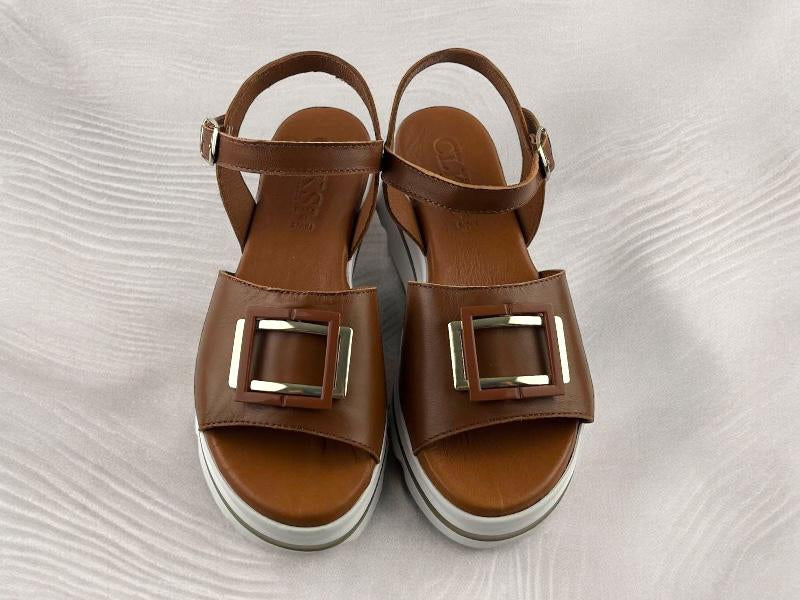Sandalias planas de mujer en color marrón avellana, con detalles geométricos metálicos en la pala de las sandalias. Vista superior de las sandalias sobre fondo de madera blanca.