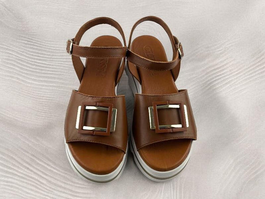 Sandalias planas de mujer en color marrón avellana, con detalles geométricos metálicos en la pala de las sandalias. Vista superior de las sandalias sobre fondo de madera blanca.