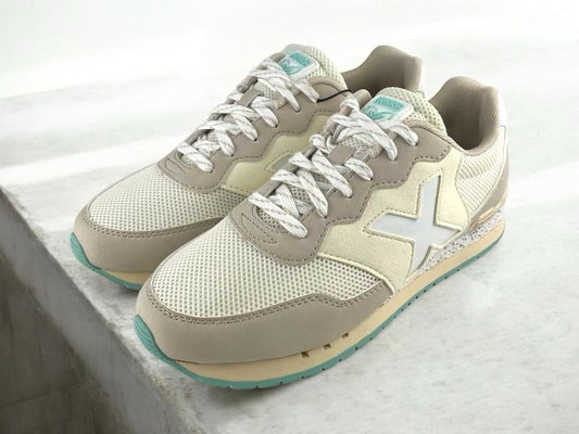 Vista lateral izquierda de unos tenis de mujer, estilo retro running, de la marca Munich en color beige claro, con malla transpirable, cordones y suela de goma sobre fondo de mármol beige.