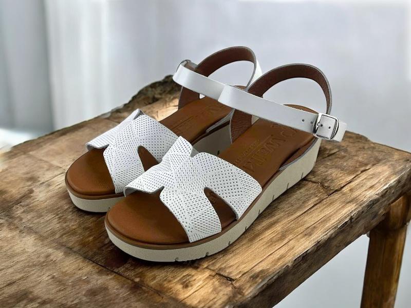 Sandalias cómodas de mujer en piel color blanco, con pala troquelada y cierre de hebilla al tobillo sobre fondo de madera rústica.