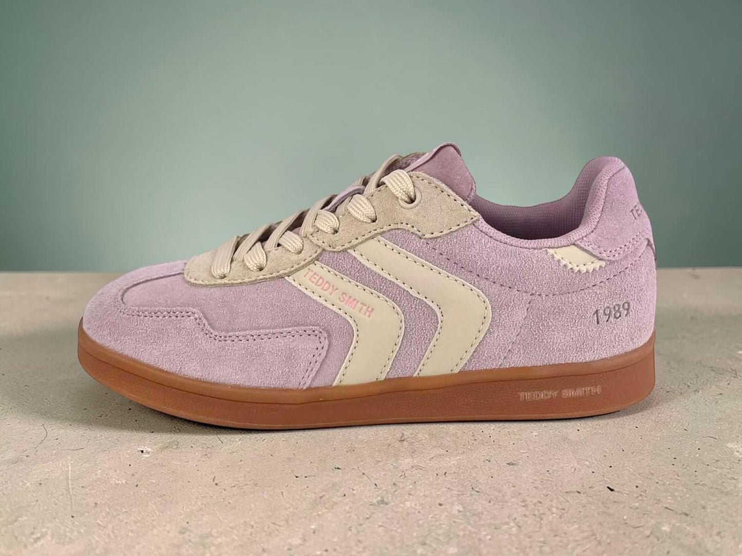 Perfil de una zapatilla de deporte Teddy Smith para mujer. Parte superior de ante rosa lavanda con rayas laterales crema y acentos beige o amarillo suave. Suela de goma color gum caramelo. Texto "TEDDY SMITH" en el lateral y "1989" en el talón. Cordones beige.