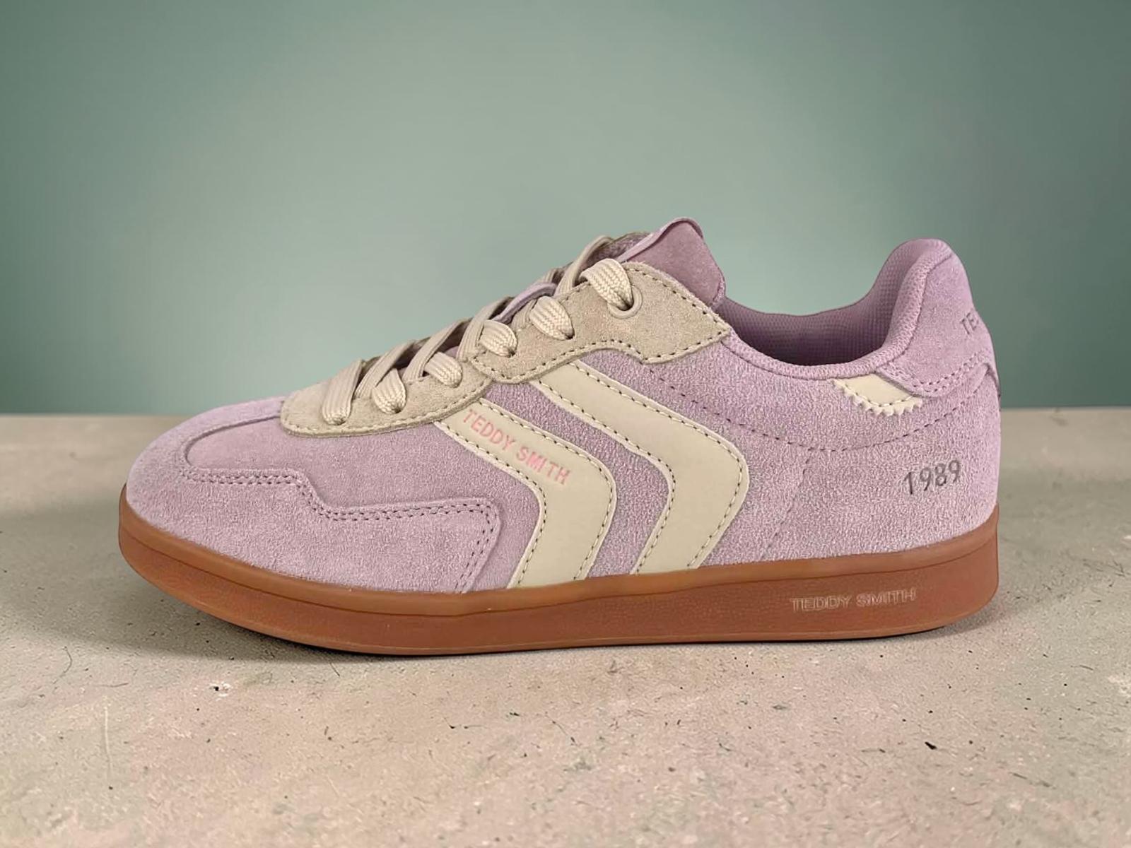 Perfil de una zapatilla de deporte Teddy Smith para mujer. Parte superior de ante rosa lavanda con rayas laterales crema y acentos beige o amarillo suave. Suela de goma color gum caramelo. Texto "TEDDY SMITH" en el lateral y "1989" en el talón. Cordones beige.